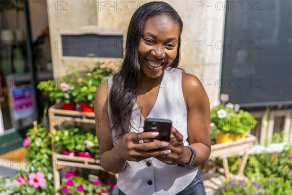 Smiling black business owner using mobile phone app managing online orders in front of her flower shop