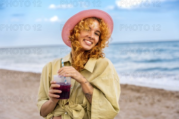 Young woman with curly red hair and pink hat enjoying a refreshing cold beverage on a sunny beach