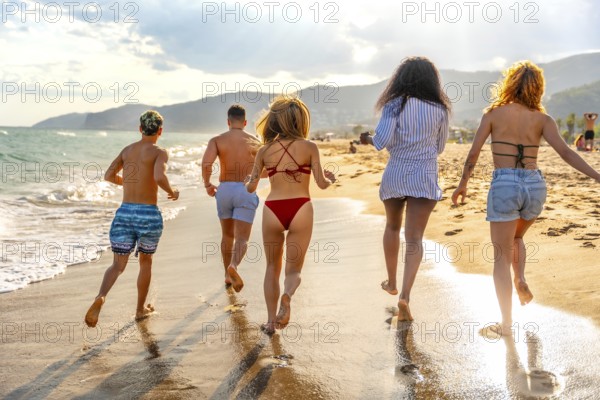 Five young friends running towards the sea waves on a sandy beach at sunset, enjoying their summer vacation