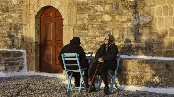 Two elderly people sitting on blue chairs in front of a stone wall and talking, Easter 2025, Church of the Dormition of the All-Holy God Bear, Kimisis tis Theotokou, Asklipio, Rhodes, Dodecanese, Greek Islands, Greece, Europe