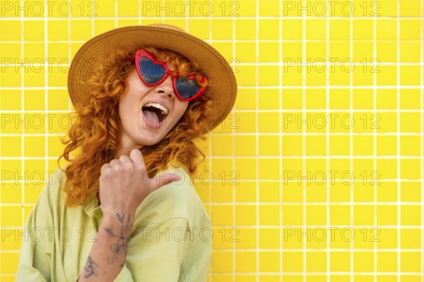 Cheerful influencer pointing her thumb while smiling, wearing a straw hat and heart shaped sunglasses on a yellow tiled wall