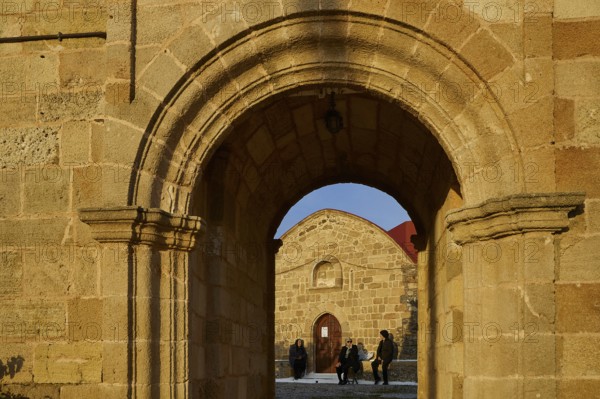 View through a stone arch onto people sitting and chatting in front of a church, Easter 2025, Church of the Dormition of the All-Holy God Bear, Kimisis tis Theotokou, Asklipio, Rhodes, Dodecanese, Greek Islands, Greece, Europe