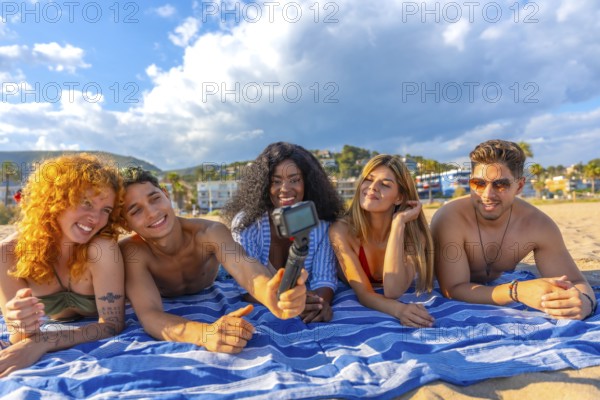 Happy multi ethnic friends lying together on a towel, capturing a joyful selfie on the beach while enjoying a sunny summer day