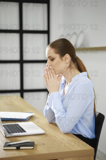 A business woman sits at her desk with a laptop, hands clasped in front of her mouth, visibly deep in thought and reflection