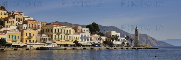 Historic coastal buildings, a striking tower and the sea under a bright blue sky, Symi Town, Symi, Dodecanese, Greek Islands, Greece, Europe