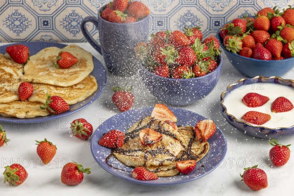 Icing sugar falls on pancakes with strawberries and chocolate syrup, fresh strawberries and yoghurt in the background