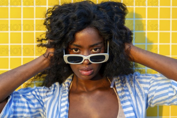 Fashionable model with curly hair touching her head while wearing white sunglasses and posing in front of a bright yellow tiled wall