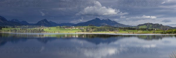 Panorama at sunrise over Lake Hopfensee to the sunlit western shore with farms, behind it receding thunderclouds, Lake Hopfensee, Ostallgäu, Allgäu, Bavaria, Germany, Europe
