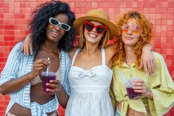 Three multi ethnic young women hugging each other and drinking juice with straw, standing against a red tiled wall