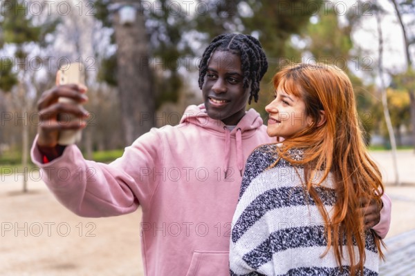 Young multiracial couple smiling together while capturing a joyful selfie with a smartphone in a vibrant park setting