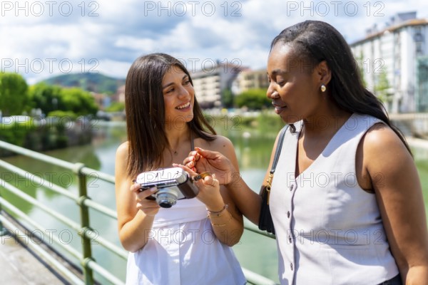 Two female friends looking at a vintage camera, standing next to a river in a sunny city, enjoying their vacation together