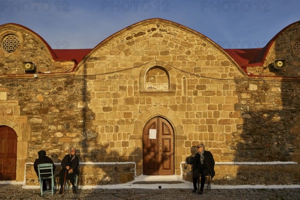 People sitting in front of a stone church in warm light and quiet surroundings, Easter 2025, Church of the Dormition of the All-Holy God Bear, Kimisis tis Theotokou, Asklipio, Rhodes, Dodecanese, Greek Islands, Greece, Europe