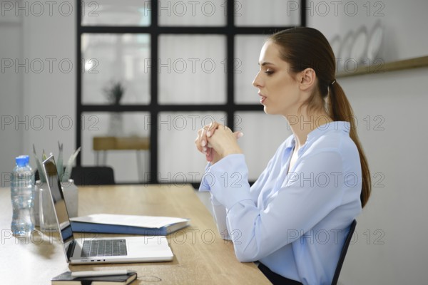 A woman wearing a light blue blouse sits at a modern desk engaged in a virtual meeting, concentrating intently as her laptop is open in front of her. The workspace is bright and organized