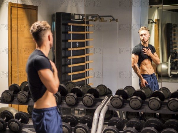 Young Shirtless Handsome Muscular Man Admiring His Muscles in Gym Mirror or Checking Progress and Training Results, as seen from Behind