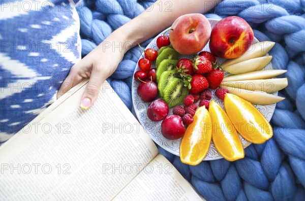Plate with fresh fruit salad on a blue plaid and book. Woman reading the book and eating fresh fruits. Close up. Top veiw. Cozy and healthy concept