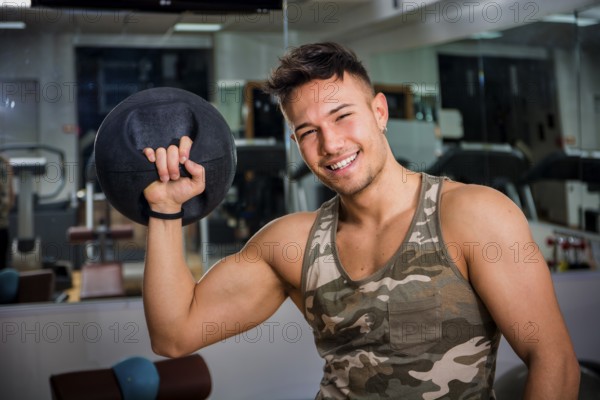 Attractive young athletic man exercising in gym, working out using kettlebell, looking at camera with a smile