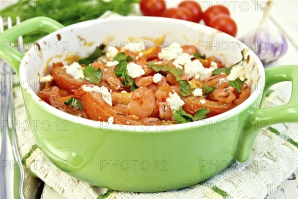 Shrimp and tomatoes baked with feta cheese in a roasting pan on a kitchen towel, parsley and garlic on a wooden boards background