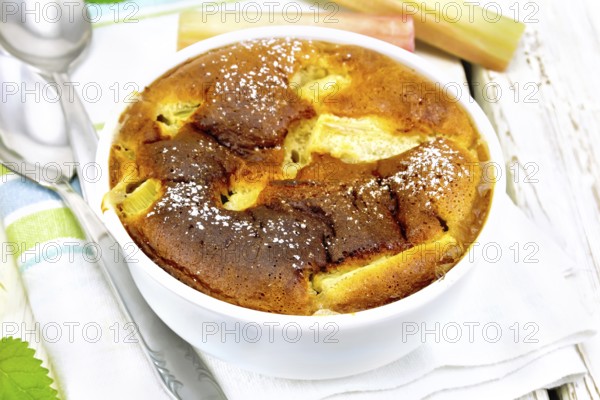Pie klafouti with rhubarb in the bowl, mint and rhubarb stalks on a towel on the background of light wooden board