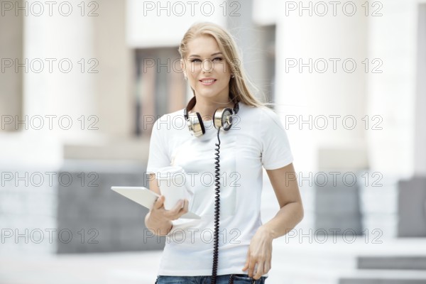Happy young woman with vintage headphones around her neck, holding a cup of coffee to go and surfing the internet on a tablet PC, against an urban background