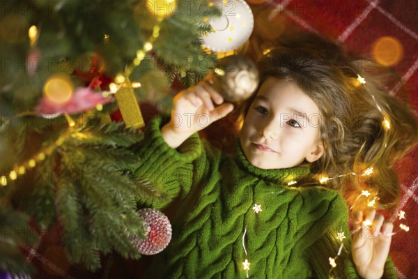 A girl with long hair and garlands lies on a red plaid under a Christmas tree with toys in a warm knitted sweater. Christmas, New Year, children's emotions, joy, expectation of a miracle and gifts