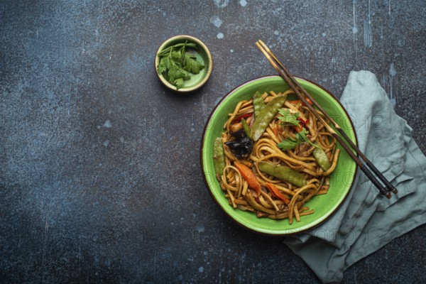 Asian stir fry udon noodles with chicken, vegetables, mushrooms in green ceramic bowl with wooden chopsticks on rustic dark blue concrete background from above copy space, Chinese or Thai fast food, food photography