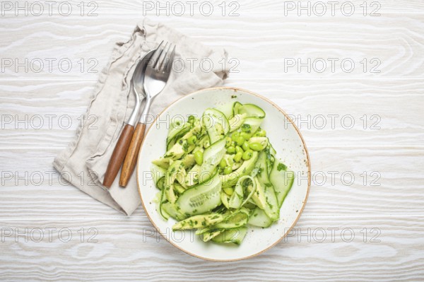 Healthy vegan green avocado salad bowl with sliced cucumbers, edamame beans, olive oil and herbs on ceramic plate top view on white wooden rustic table background, food photography, food photography