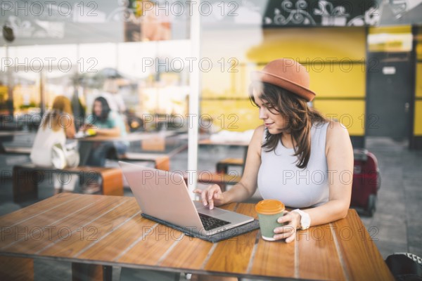 Beautiful young caucasian woman sitting on a terrace in a cafe in summer at a wooden table in a hat and a plate uses technology, working behind a laptop booking hotels trip in the summer