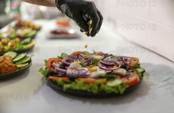 Chef wearing black gloves adding corn grains to a greek salad with tomatoes, red onions, olives, cucumbers, feta cheese and lettuce on a black plate in an indian restaurant kitchen
