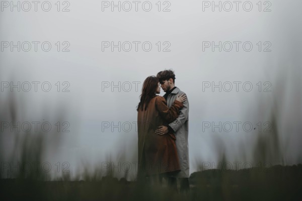 Couple holds each other in a moody open landscape, dressed in trench coats