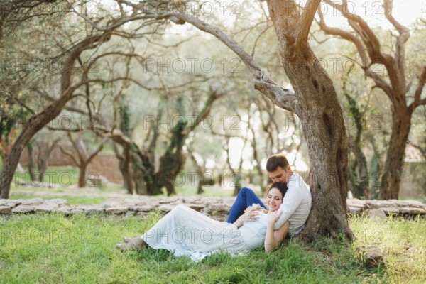 Kotor, Montenegro - 21.06.17: Seated man hugs a woman lying next to him on green grass and kisses her on the crown near a tree in an olive grove