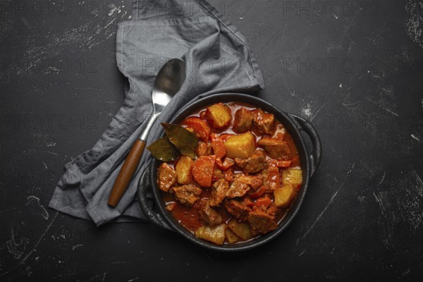 Beef meat stew with potatoes, carrot and delicious gravy in black casserole pot with bay leaves with spoon on black dark rustic concrete background from above, food photography