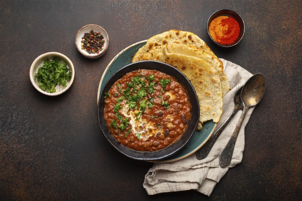Traditional Indian Punjabi dish Dal makhani with lentils and beans in black bowl served with naan flat bread, fresh cilantro and two spoons on brown concrete rustic table top view, food photography, food photography