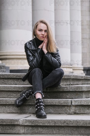 Portrait of young blonde woman with red lips wearing leather jacket on corporate building stairs. Career businesswoman. Stylish female biker. Urban street city lifestyle