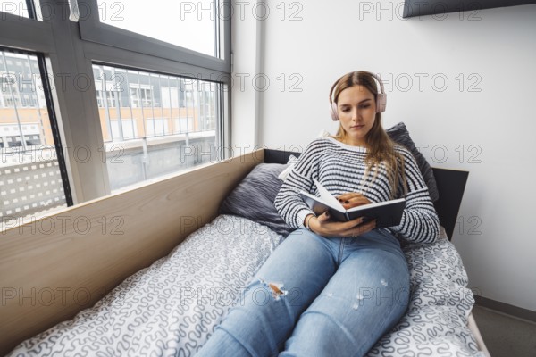 Blonde young caucasian woman lying on her dorm room bed next to a window putting on headphones, listening to music on a cold cloudy autumn day