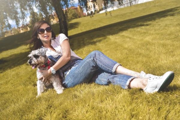 Pretty adult caucasian happy woman resting in the park on a sunny day with her beloved dog. Female lay on the grass smiling and looking at the camera
