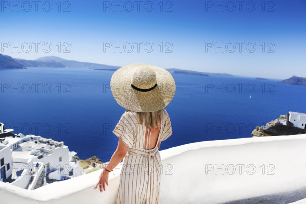 Young woman in a white dress and straw hat, walking at the city of Oia, island of Santorini, Greece. Concept of leisure, tourism, travel, vacations, romance, love