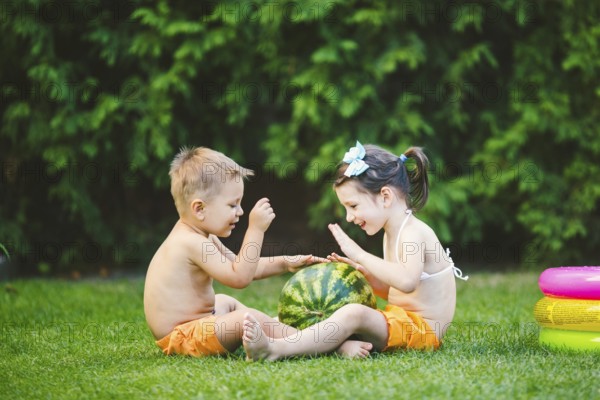 Two children, Caucasian brother and sister, sitting on green grass in backyard of house and hugging big tasty sweet watermelon berry, green striped colour. Theme healthy food and ecological product