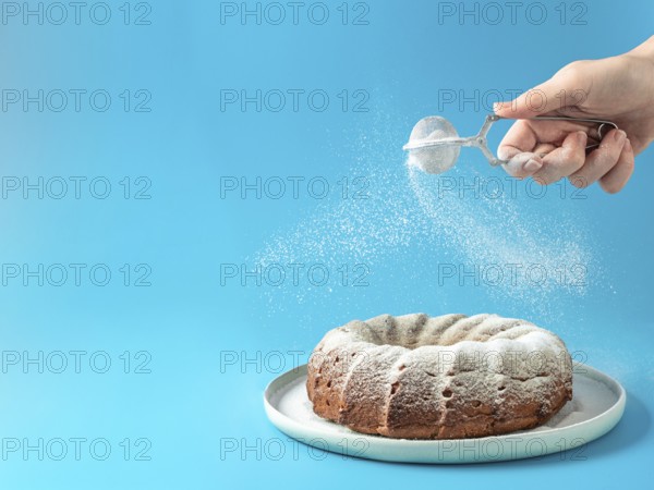 Woman's hand sprinkling icing sugar over fresh home made bundt cake. Powder sugar falls on fresh perfect bunt cake over blue background. Copy space for text. Ideas and recipes for breakfast or dessert