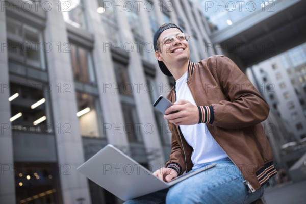 Happy handsome student looking at computer screen while typing something on laptop keyboard during work break outdoors, teenager looking at the notebook sitting at street