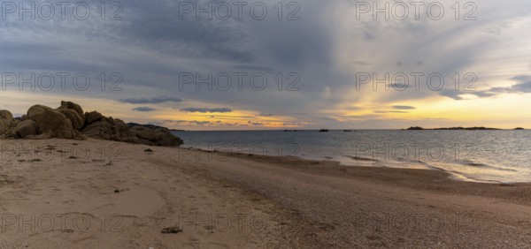 A panorama view of a golden sand beach at sunset with calm ocean water and red granite rocks