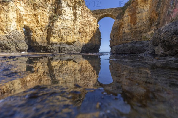 Yellow rocks and cliffs on a sandy beach. A bay by the sea, over which the old stone bridge of the Pinhão fortress connects two cliffs. Praia da Caldeira, Lagos. Algarve, Portugal, Europe
