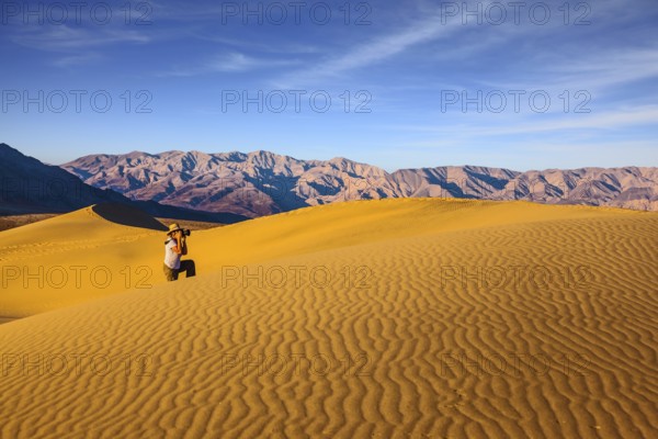 Woman in striped shirt and straw hat photographing sand waves. Bright morning in picturesque part of Death Valley. Mesquite Flat Sand Dunes