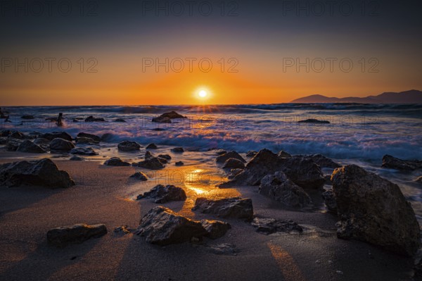 Sunset with rocks in the foreground on the beach. The sea is moving and the sky glows in shades of orange and purple, Mastichari, Kos Island, Greece, Europe
