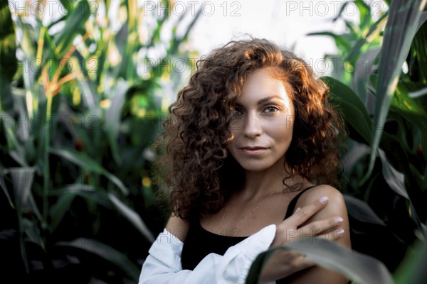 A confident woman with curly hair stands among corn stalks, crossing her arms