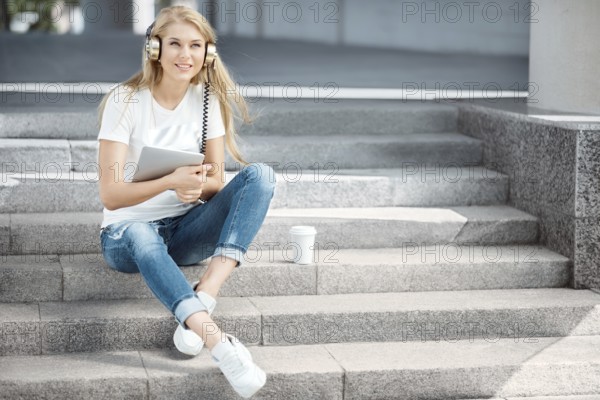 Happy young woman with vintage headphones and a cup of coffee to go, surfing the internet on a tablet PC, listening to music and sitting on a staircase in front of an urban background