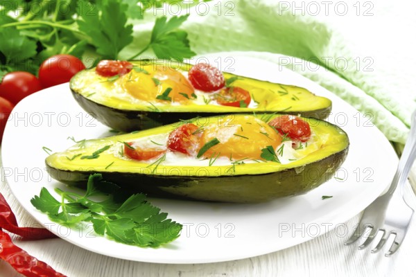 Scrambled eggs with cherry tomatoes in two halves of avocado in a plate, green towel and fork on the background of white wooden board