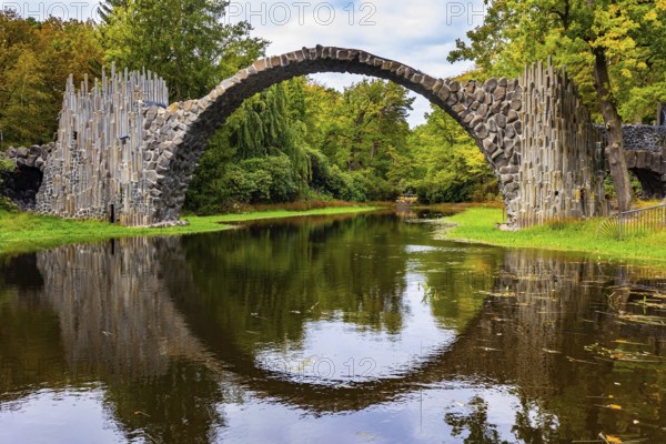 The Devil's Bridge on the Rakots River. The basalt columns of the bridge form a perfect circle with their reflection in the water. Germany. Picturesque narrow bridge in the park Kromlau