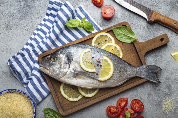 Raw fish dorado with ingredients lemon, fresh basil, cut cherry tomatoes, uncooked rice on wooden cutting board with knife on rustic stone background top view, cooking healthy fish dorado concept, food photography, food photography
