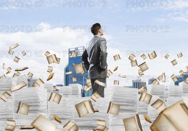 Confident businessman in suit standing on pile of documents among flying books with cityscape on background. Mixed media