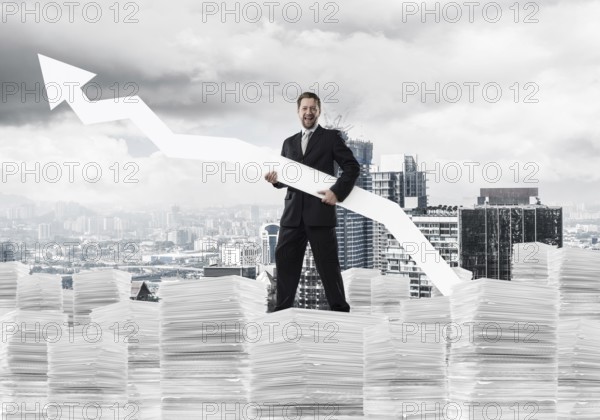 Young and ambitious businessman in suit holding big white arrow in hand and pointing away while standing on paper column